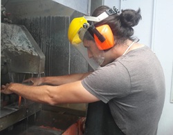 A man wearing a gray shirt and protective equipment holds a rock up to an industrial rock saw.